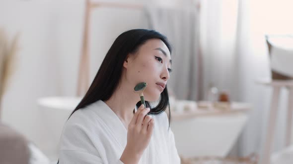 Young Attractive Asian Woman Making Self Massage with Natural Stone Massager Sitting at Bathroom at alt