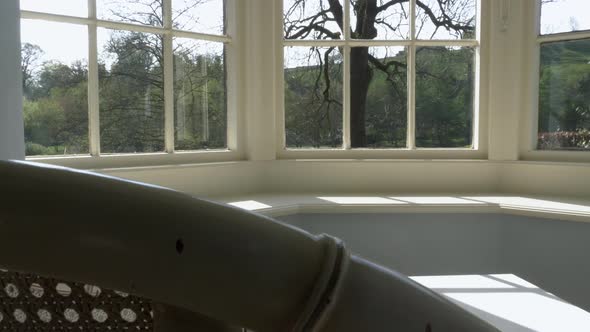 Slow slide left over top of empty white wicker chair in window bay overlooking garden alt