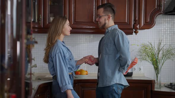 Side View of Joyful Young Man Surprising Woman with Gift on Weekend Morning in Kitchen alt