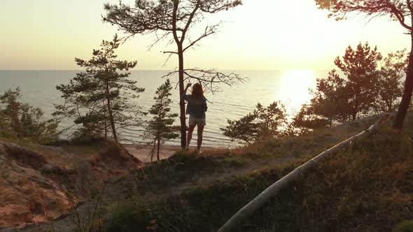 Young Woman Enjoys Beautiful Scenic Sunset at Baltic Sea Standing at Tree on Hill. Slow Aerial Orbit alt