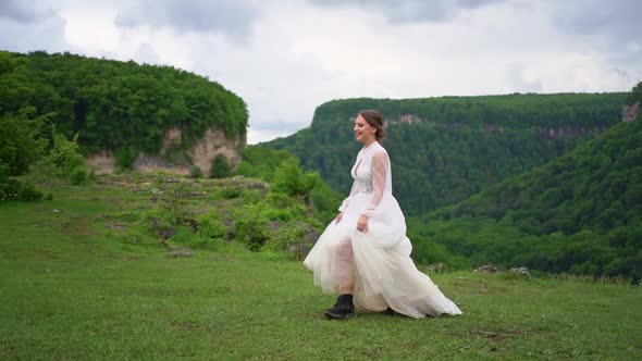 A Girl in a Wedding Dress and Black Boots Goes on the Lawn in the Mountains alt