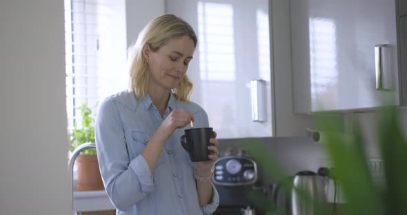 Blond woman standing stirring in coffee cup alt