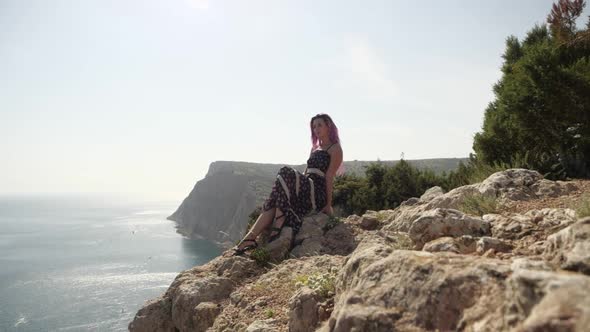 Slender Girl in a Long Dress with Pink Hair Sits on the Edge of the Cliff