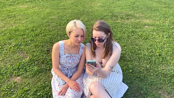 Two Girls in the Park Use a Smartphone and Discuss alt
