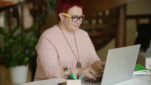 Modern middle-aged woman in home office at laptop typing text smiling Non-binary