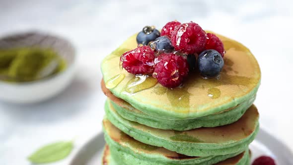 Pouring honey on green matcha pancakes with berries on a black plate. alt