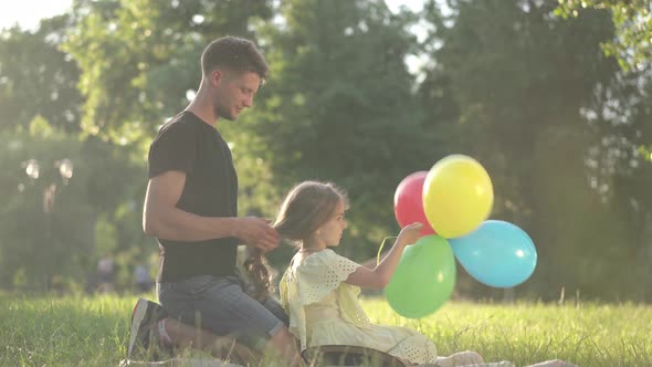 Side View Relaxed Daughter Playing with Colorful Balloons As Young Father Braiding Hair Sitting on alt