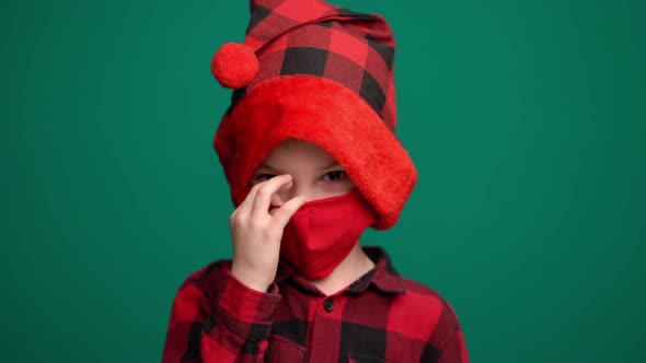 Portrait of Little Boy in Santa Hat Wears a Red Medical Mask, Looking at Camera alt