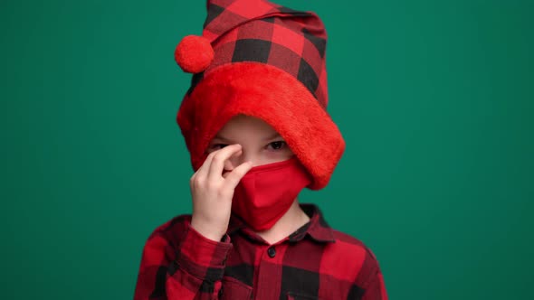 Portrait of Little Boy in Santa Hat Wears a Red Medical Mask, Looking at Camera