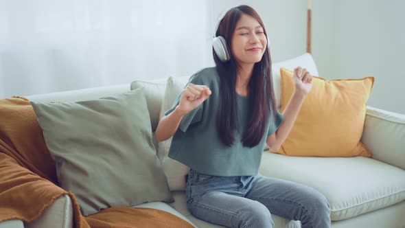 Woman wear wireless headphones having fun listen music in smartphone and sit on sofa in living room alt