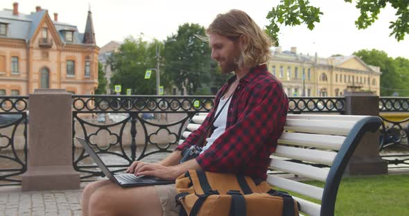 Side View of Traveler Man Wearing Casual Clothes Using Laptop Sitting on Bench Outdoors in Street alt