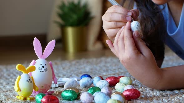 A girl draws a pattern on an Easter egg next to an egg in the shape of a rabbit