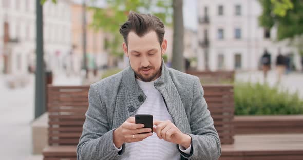 Young Man with Mustaches and a Beard Sitting on a Bench in the Square Scrolling the Smartphone alt
