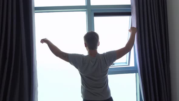 Back of View of Young Man Unveil Curtain and Stretches Arms in the Morning