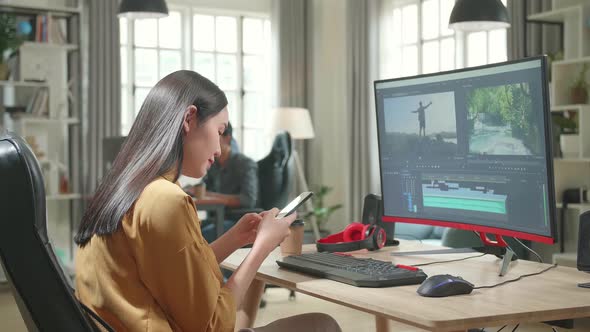Asian Female Video Editor Works And Using Mobile Phone With Her Personal Computer In Office alt