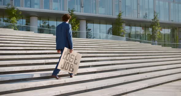 Young Man in Suit Holding Lost Job Cardboard Sign Walking Outside Business Center alt