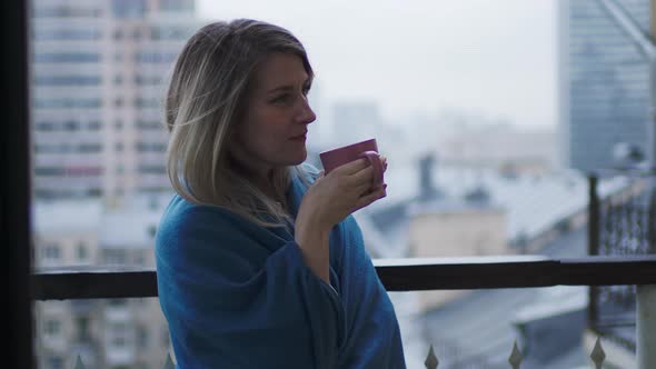 A Young Beautiful Woman with a Mug in Her Hands Stands on the Balcony alt