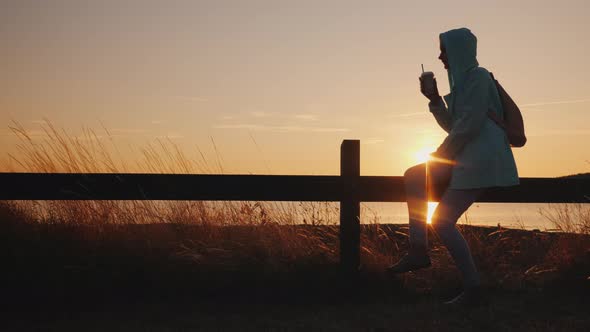A Woman Sits in a Picturesque Rural Place on the Fence Drinks Coffee alt
