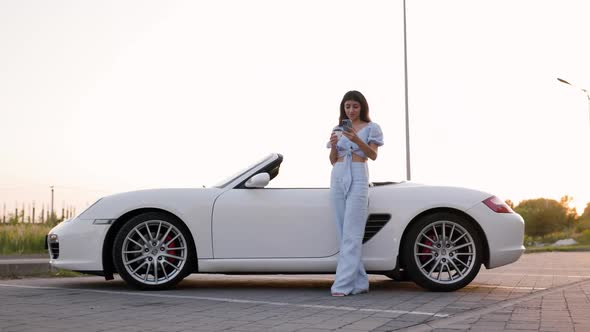 Beautiful Stylish Woman Leans on Her Luxury White Convertible and Drinks Cup of Coffee alt