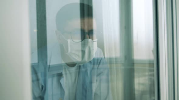 A Man in a Protection Mask Is Looking Outside of the Window alt