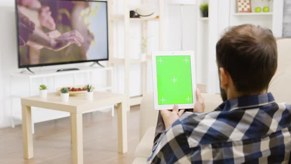 Man Looking at a Movie on the TV While Holding a Green Screen Digital Tablet PC alt