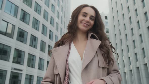 Smiling girl with brown hair stands at the corporate building for a break, dressed in an elegant alt