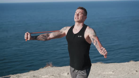 Tattooed Sportsman Does Exercises with Elastic Bands with Ocean View alt