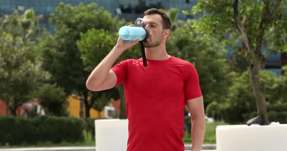 Muscular man wearing red sports shirt walking in park and breathing hard, drinking water from bottle