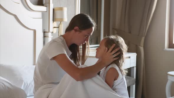 Young Mother Caresses Her Little Daughter in Bedroom at Home alt