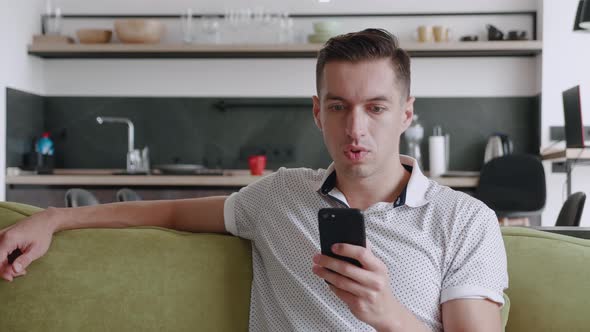 Shocked Man Reacting Message on Smartphone Covers His Mouth in Fright Sitting on Sofa at Home Living alt