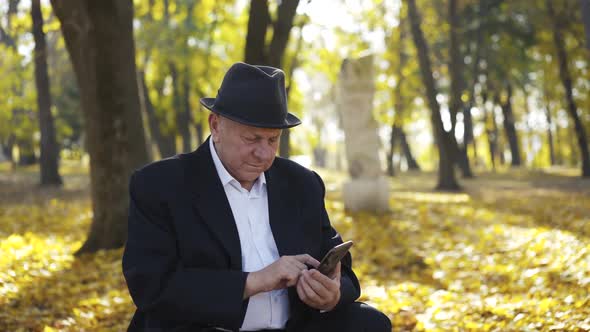 Joyful Senior Man in Suit and Hat Uses Smartphone and Talks with Smile in Park alt