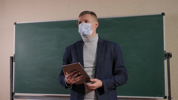 Young Male Teacher in a Medical Mask on His Face at School Near the Blackboard alt