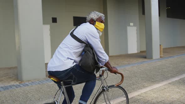 African american senior man wearing face mask riding bicycle in corporate park alt