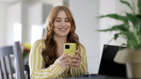 Smiling woman writing text message on smartphone while sitting at desk