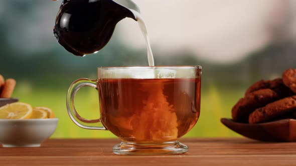 Pouring Milk Into Black Tea in Glass Cup on a Wooden Table alt