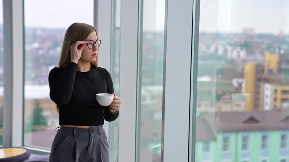 Beautiful female with cup of coffee standing by the window alt