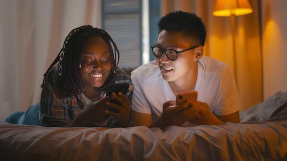 Multiracial Young Couple Using Mobile Phones Relaxing in Bed in Evening alt