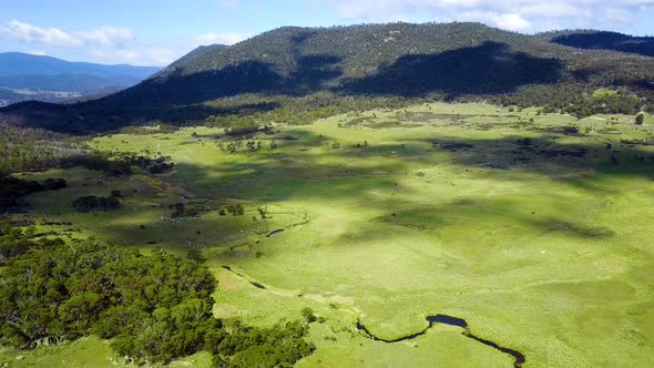 Drone footage of a lush green valley with a winding river and moving cloud shadows and mountains in alt