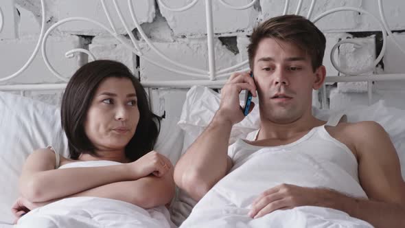 Young Couple Lying in Bed and Man Cheating on Woman Chatting on Smartphone alt