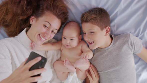 Happy Mother with Her Kids Are Making a Selfie or Video Call To Father or Relatives in a Bed alt