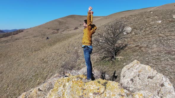 Man Stands on a Stone on a Hillside with His Hands Up and Enjoys Freedom Shooting Drone