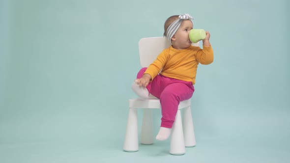 Toddler 12-17 months old sits on a small white chair and drinks yogurt from a baby cup alt