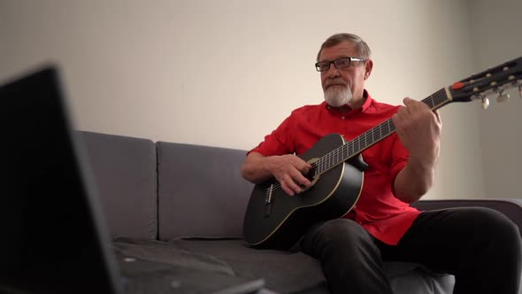 Mature Man Learns To Play the Guitar Online While Sitting on the Couch Near the Laptop alt