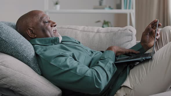 Senior Older Mature African Man Typing on Laptop Lying on Sofa at Home Office Browsing Internet alt
