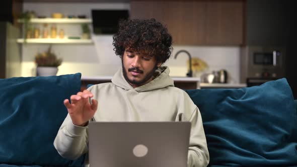 Cheerful Indian Man Wearing Casual Hoodie Sitting on the Sofa and Using Laptop at Home