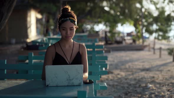Young influencer girl working on laptop at beach lit by sunset, Bali alt