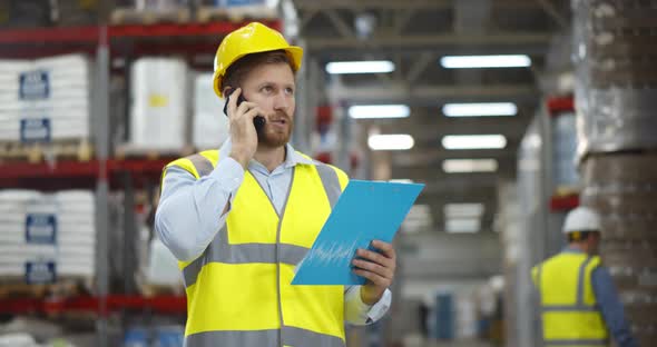Male Worker Looking at Clipboard While Talking on Mobile Phone in Warehouse alt