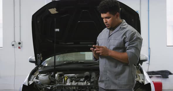 African American male car mechanic looking at an open car engine and talking on a smartphone alt