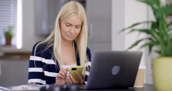 Blond woman using smartphone at desk with laptop