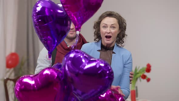 Carefree Cheerful Adult Loving Couple Waving Looking at Camera with Heart Shape Balloons at Front alt